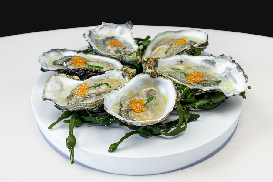 Six oysters on the half shell, garnished with orange roe and a light sauce, are arranged in a circle on a bed of green seaweed atop a white round plate.