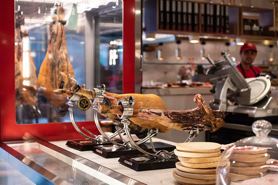 A leg of cured ham is mounted on a stand at a deli counter. Wooden plates are stacked nearby, and a worker in red stands by a meat slicer in the background, surrounded by shelves of bottles.