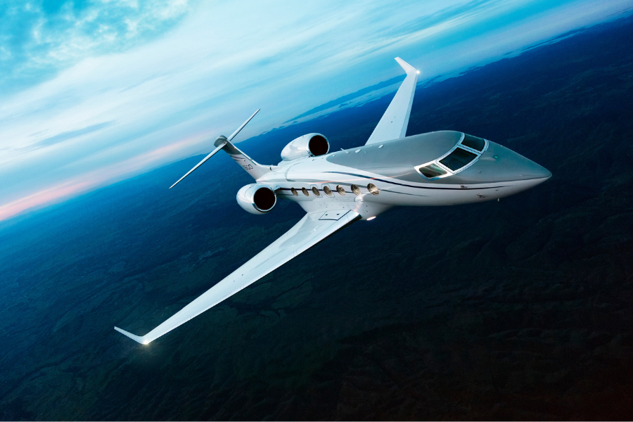 A sleek white private jet flies high above a landscape of hills and forests under a blue sky with scattered clouds during sunset or sunrise.