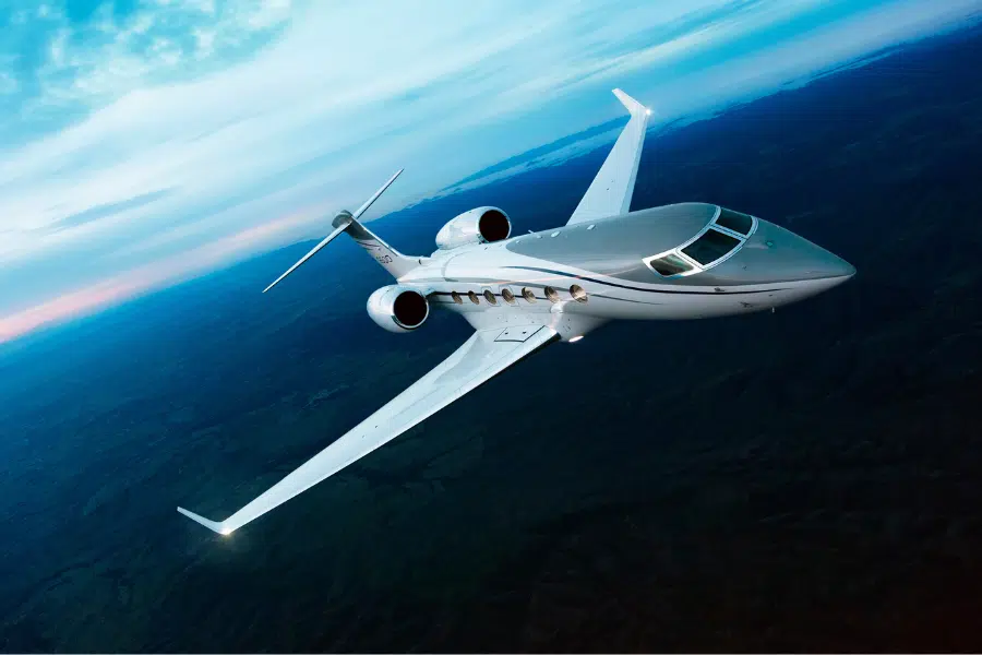 A sleek white private jet flies high above a landscape of hills and forests under a blue sky with scattered clouds during sunset or sunrise.