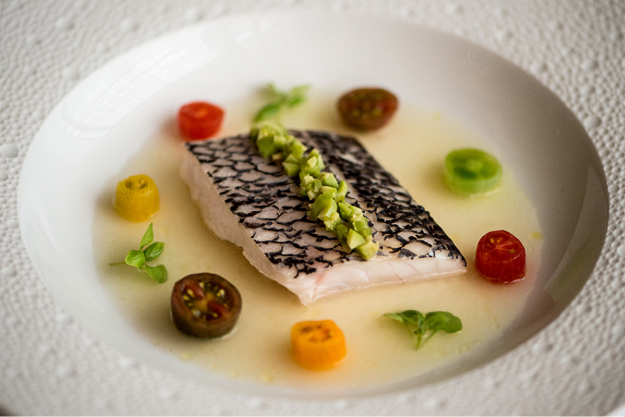 A plated dish featuring a fillet of fish with black skin, topped with diced green garnish, surrounded by colorful cherry tomato halves and small green herbs in a light sauce.