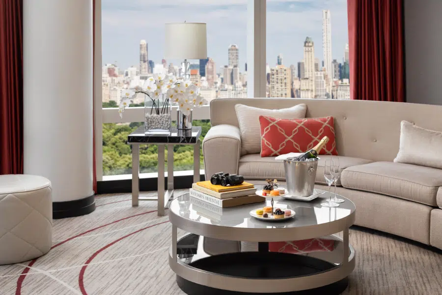 A modern hotel suite with a beige sofa, a glass coffee table set with desserts, a champagne bucket, books, and flowers. Large windows reveal a city skyline and a park outside. Red curtains frame the view.