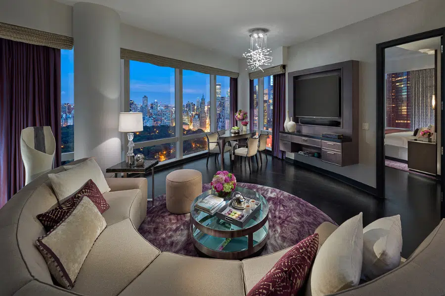A modern, luxurious hotel suite with a curved sofa, glass coffee table, and large windows offering a stunning cityscape view at dusk. The room is softly lit, with purple accents and a separate bedroom visible.