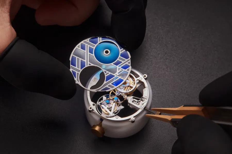 A close-up of gloved hands assembling a luxury mechanical watch, placing a decorative blue and silver watch face over intricate gears with tweezers on a dark surface.