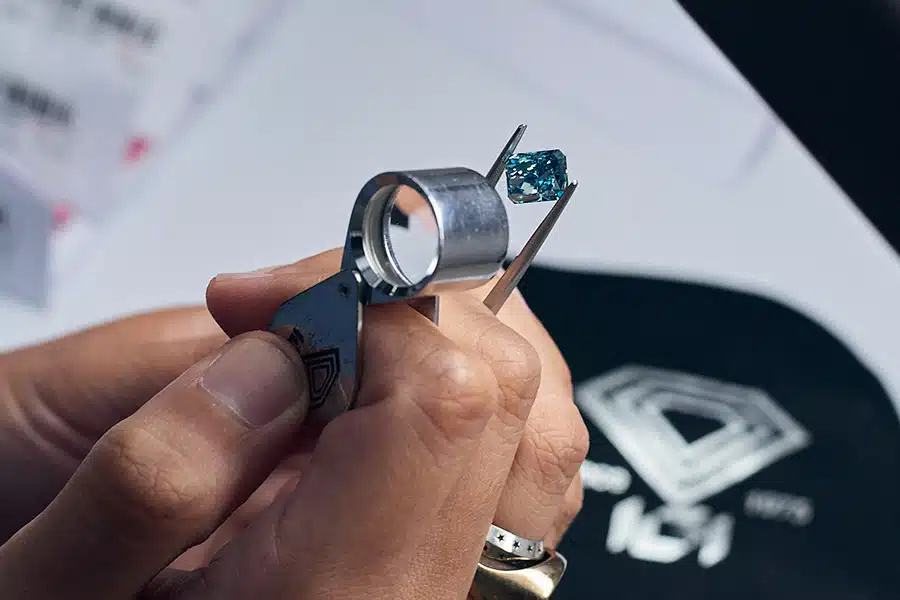 A person examines a blue gemstone with a jewelers loupe and tweezers, focusing on its details. A black cloth with a geometric logo is visible in the background.