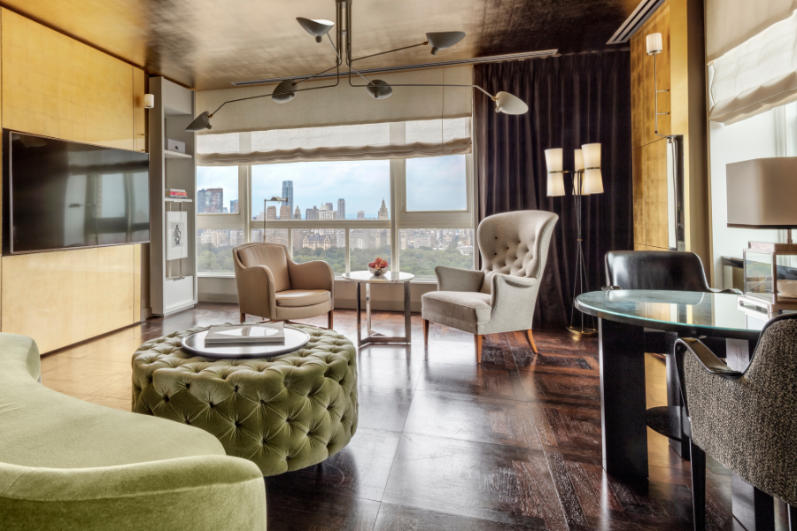 Modern living room with large windows, city skyline view, contemporary furniture including a green velvet ottoman, gray armchairs, round glass table, and wall-mounted TV; dark wood floors and neutral tones throughout.