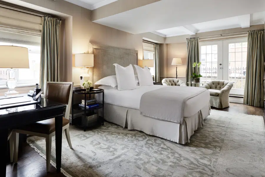A spacious, elegant bedroom with a large bed, neutral bedding, armchairs, desk, lamps, and large windows with curtains, letting in natural light onto a patterned rug and wooden floor.