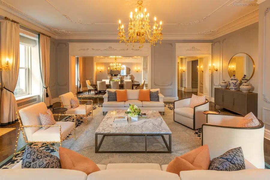 Elegant living room with beige armchairs, peach and patterned pillows, a marble coffee table, and a chandelier. Large windows with curtains and a view into a formal dining area with another chandelier.