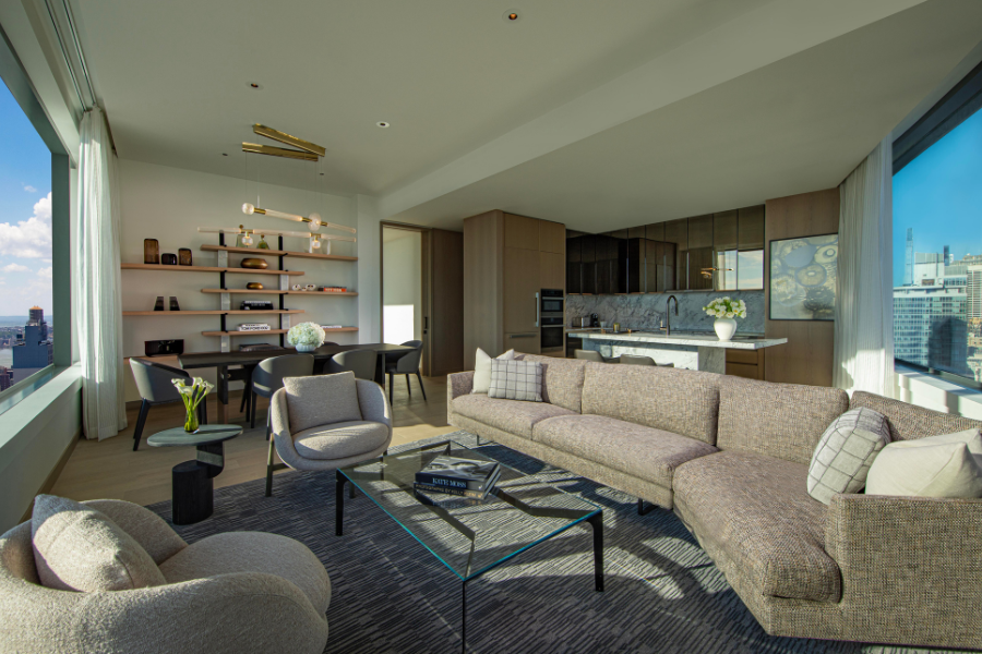 Modern, sunlit apartment living room with floor-to-ceiling windows, neutral-toned furniture, a glass coffee table, open shelving, dining area with a round table, and a sleek kitchen in the background. City skyline visible outside.