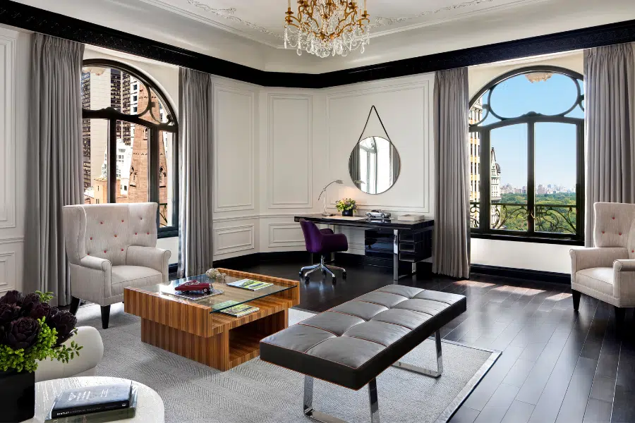 Elegant home office with large arched windows, a chandelier, a black desk, two armchairs, a modern coffee table, a bench, and cityscape views outside. Light walls contrast with dark floors and furnishings.