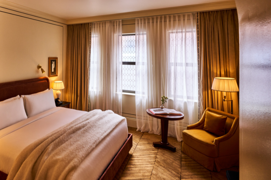 A cozy hotel room with a large bed, soft beige bedding, a wooden table, a gold armchair, two lamps, and tall windows with sheer white curtains and golden drapes, allowing warm natural light to fill the space.