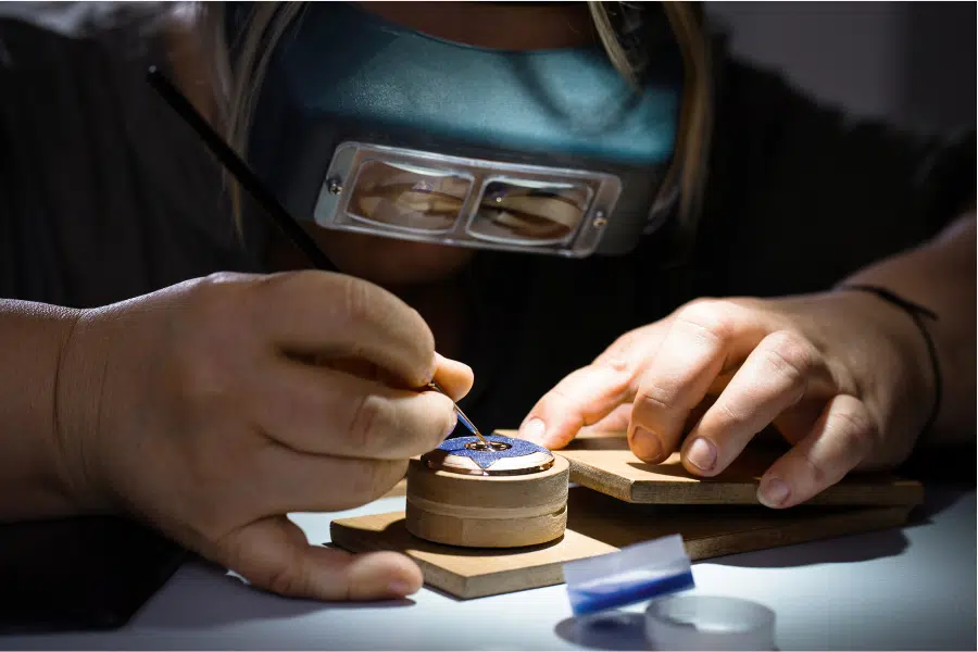A person wearing a magnifying visor uses a fine brush to paint intricate details on a small circular object, possibly a watch face or piece of jewelry, held steady by wooden supports.