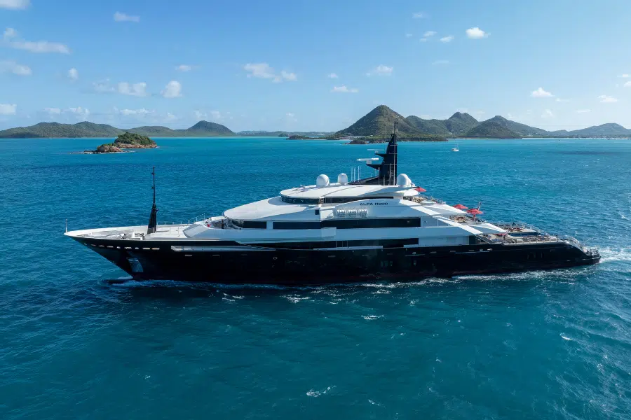 A large luxury yacht cruises on bright blue tropical waters near green, hilly islands under a clear, sunny sky.