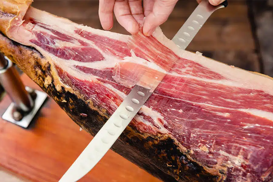 A close-up of a hand slicing thin pieces of cured ham with a long knife from a whole leg of ham mounted on a wooden stand.