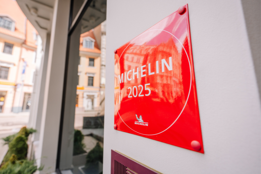 A bright red 2025 Michelin plaque is displayed on the exterior wall of a building, with a blurred city street and buildings visible in the background.