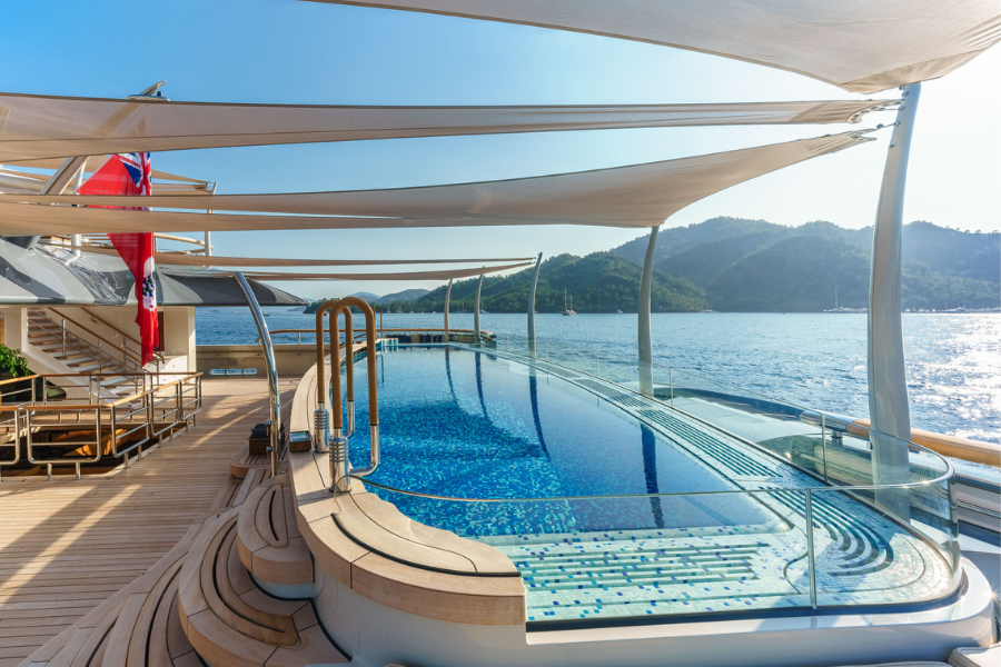 Luxury yacht deck with a curved swimming pool, shaded by modern canopies, overlooking calm blue water and forested hills in the background on a sunny day.