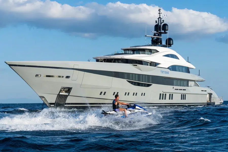 A person rides a jet ski in the foreground, creating waves on the water, with a large, modern luxury yacht in the background under a partly cloudy sky.