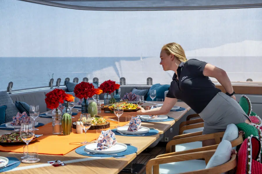A woman sets a colorful, elegantly arranged dining table on a boat. The table features plates, napkins, red flower centerpieces, and dishes of food, with the sea visible in the background.