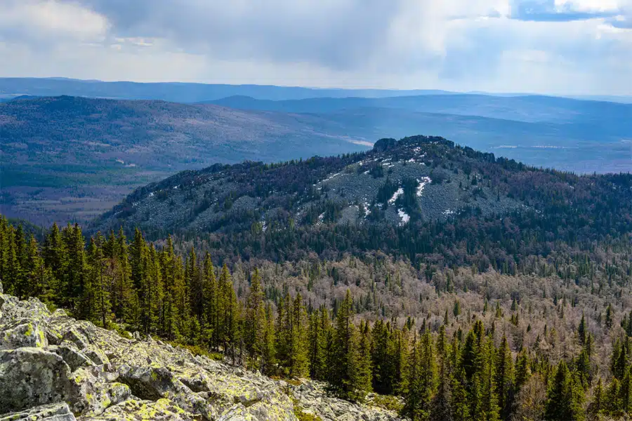 A rocky hilltop covered with patches of snow rises above dense evergreen forests, with rolling hills and a cloudy sky in the background.