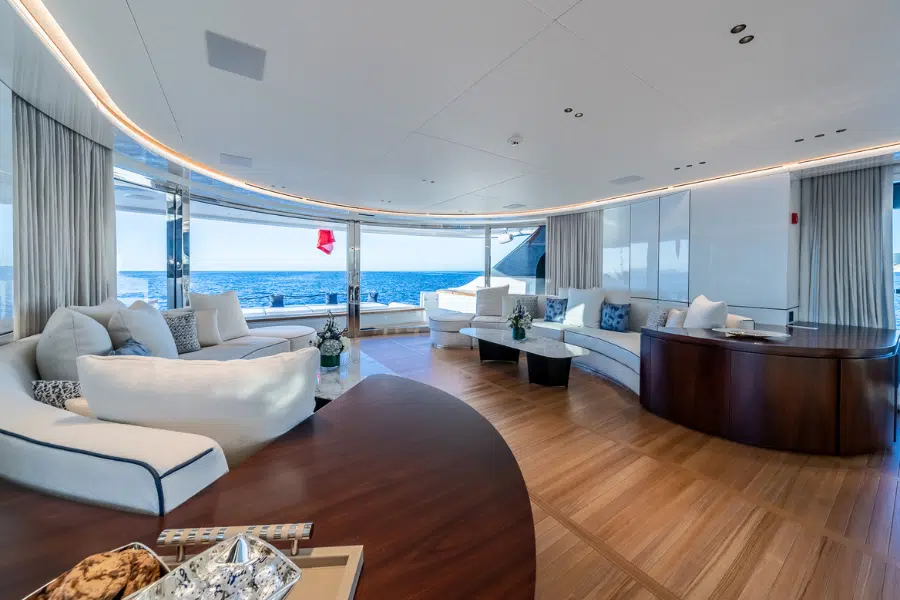Spacious, modern yacht interior with curved sofas, plush cushions, wooden floors, and large windows offering a panoramic ocean view under bright daylight.