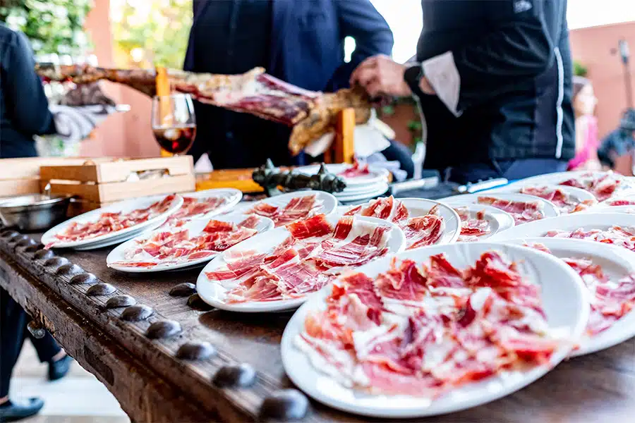 Several white plates filled with thinly sliced cured ham are arranged on a wooden table. In the background, a person slices a large leg of ham, and a glass of red wine is visible.