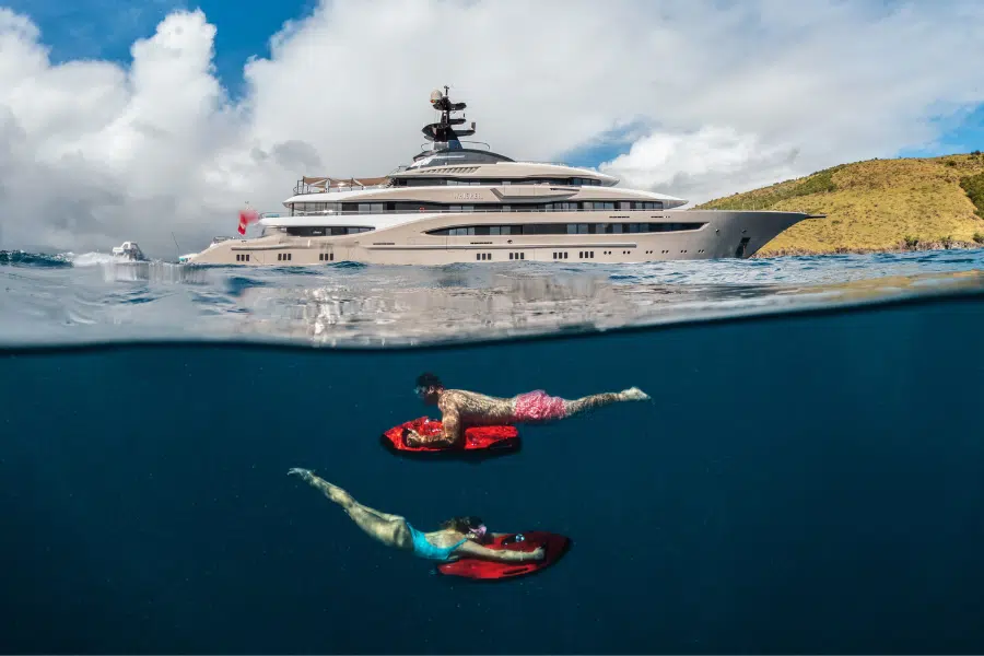 Two people ride underwater scooters in clear blue water near a large luxury yacht, with a scenic coastline and cloudy sky visible in the background above the waterline.