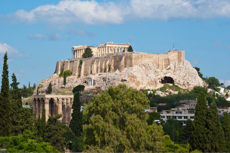 The Acropolis of Athens, featuring the Parthenon atop a rocky hill, is surrounded by trees and ancient ruins under a blue sky with scattered clouds.