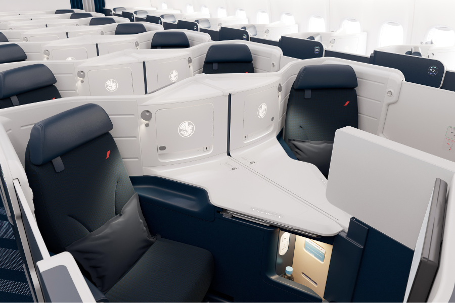 Business class seats on an airplane, arranged in a staggered layout with individual pods, dark blue and white color scheme, each seat featuring a pillow, side table, and privacy dividers.