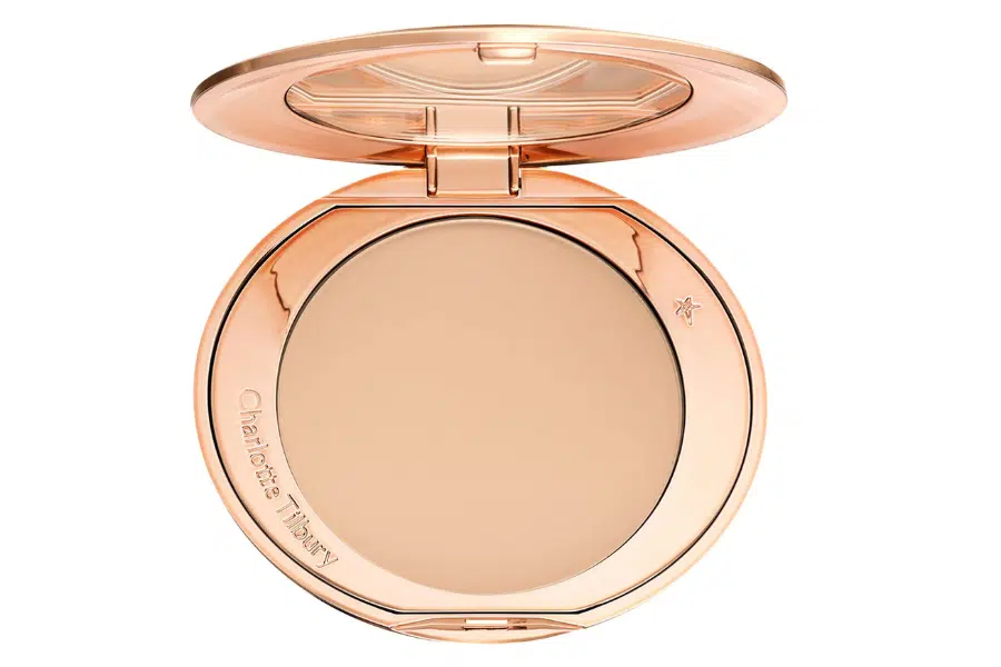 A gold compact case with a mirror, opened to reveal pressed powder makeup. The lid is reflective and engraved with “Charlotte Tilbury” along the edge.