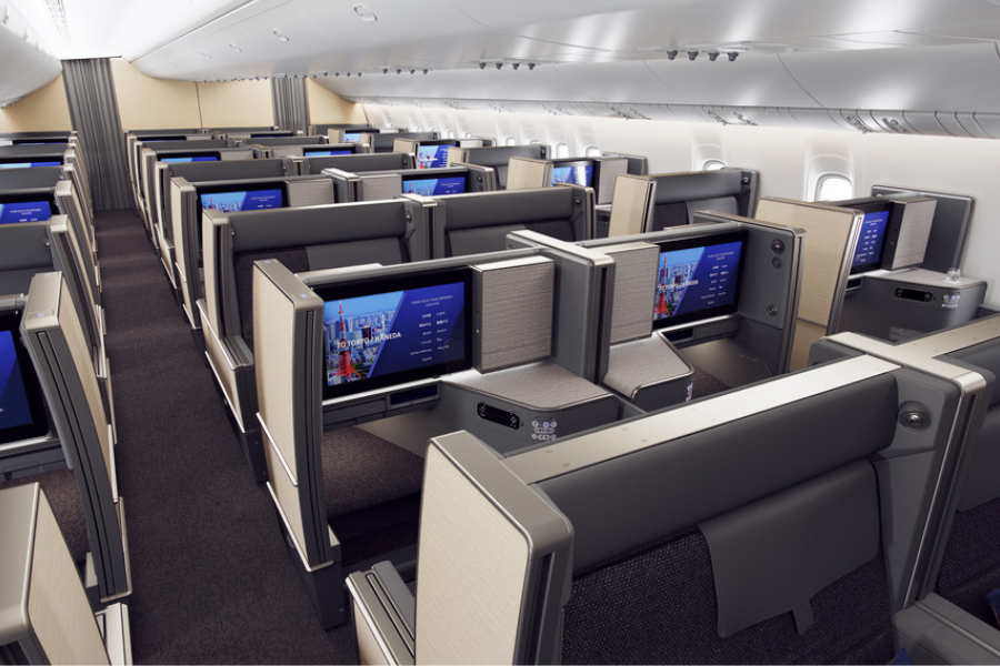 Rows of modern business class airplane seats with large personal screens, high partitions for privacy, and neutral-colored upholstery, set in a spacious and well-lit cabin interior.