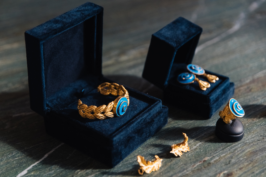 Gold jewelry set with blue accents, including a bracelet, earrings, and ring, displayed in dark blue velvet boxes on a wooden surface. Two loose gold earring backs are placed nearby.