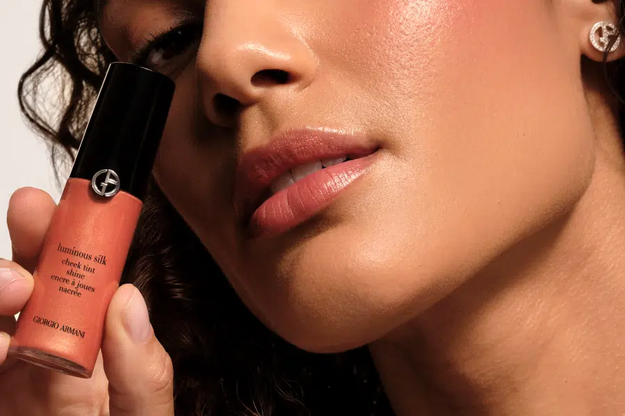 A close-up of a woman’s face with glowing skin, natural makeup, and glossy lips, holding a peach-colored Giorgio Armani Luminous Silk cheek tint near her cheek.
