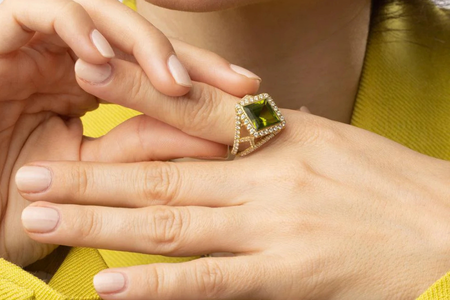 A close-up of a hand wearing a gold ring with a large green gemstone surrounded by small diamonds. The person has neatly manicured nails and is wearing a yellow garment.