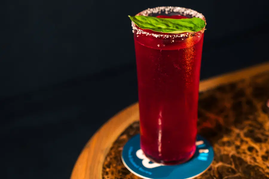 A tall glass of bright red cocktail with a salted rim and a green basil leaf garnish, placed on a blue coaster on a brown marble table.
