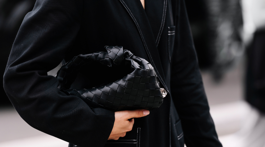 A person in a black outfit holds a black woven leather handbag with a knot detail, shown from the shoulders down in an outdoor setting.