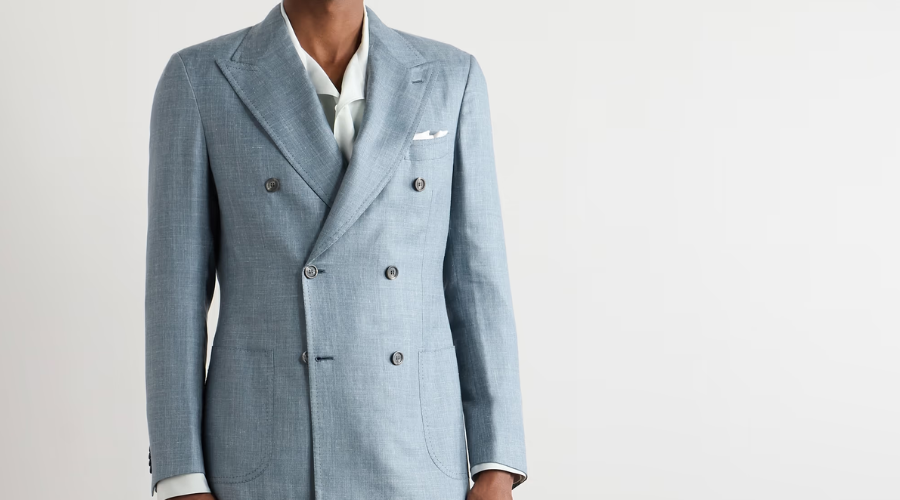 A person wearing a tailored light blue double-breasted suit jacket, white shirt, and white pocket square, standing against a plain white background. Their head is cropped out of the frame.