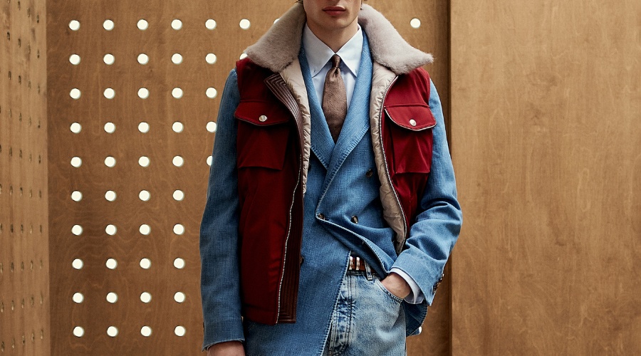 A person dressed in a blue double-breasted suit, tie, denim jeans, and a red vest with a shearling collar stands in front of a wooden wall with evenly spaced circular holes.