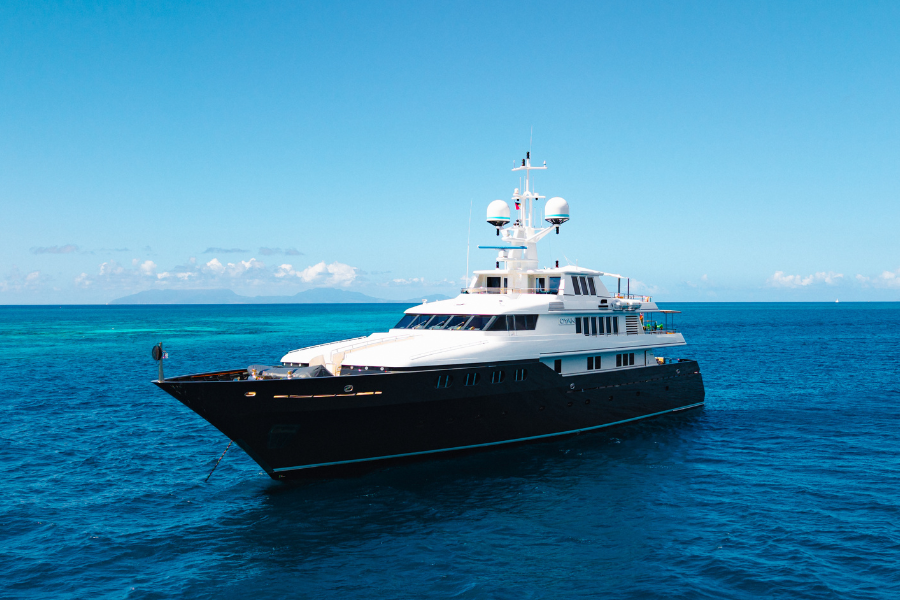 A large luxury yacht with a black hull and white upper decks floats on bright blue ocean water under a clear sky, with distant land visible on the horizon.