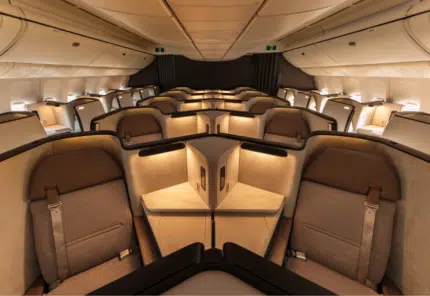 Spacious first-class airplane cabin with wide, beige and brown seats arranged in a 1-2-1 configuration, each with privacy partitions, soft lighting, and large personal space.