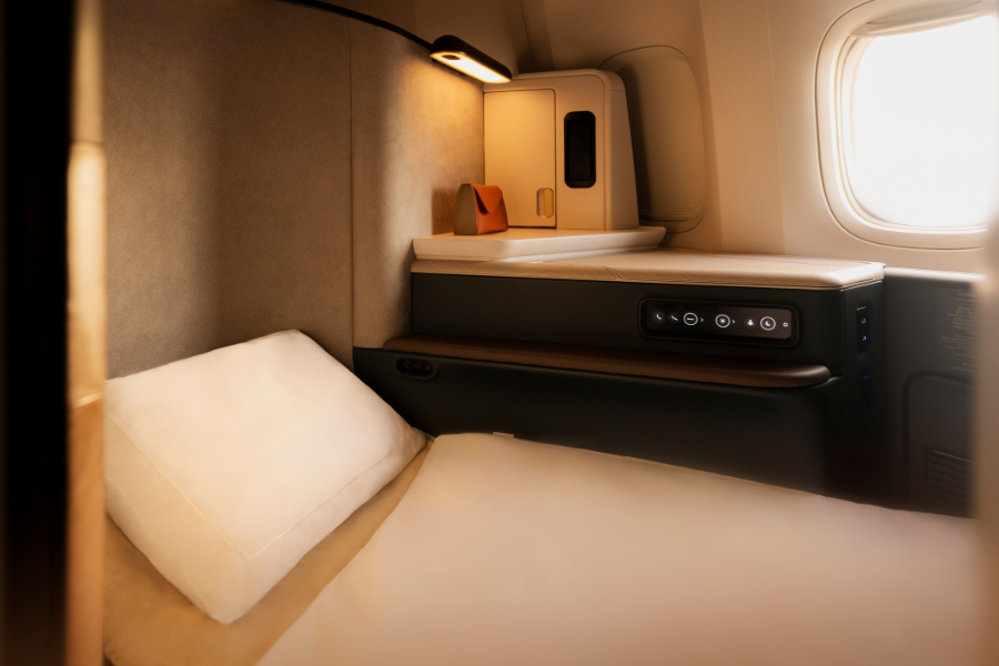 A luxurious first-class airplane suite features a neatly made bed with white bedding, a soft pillow, built-in control panel, reading light, side table, and a window allowing natural light into the cozy space.