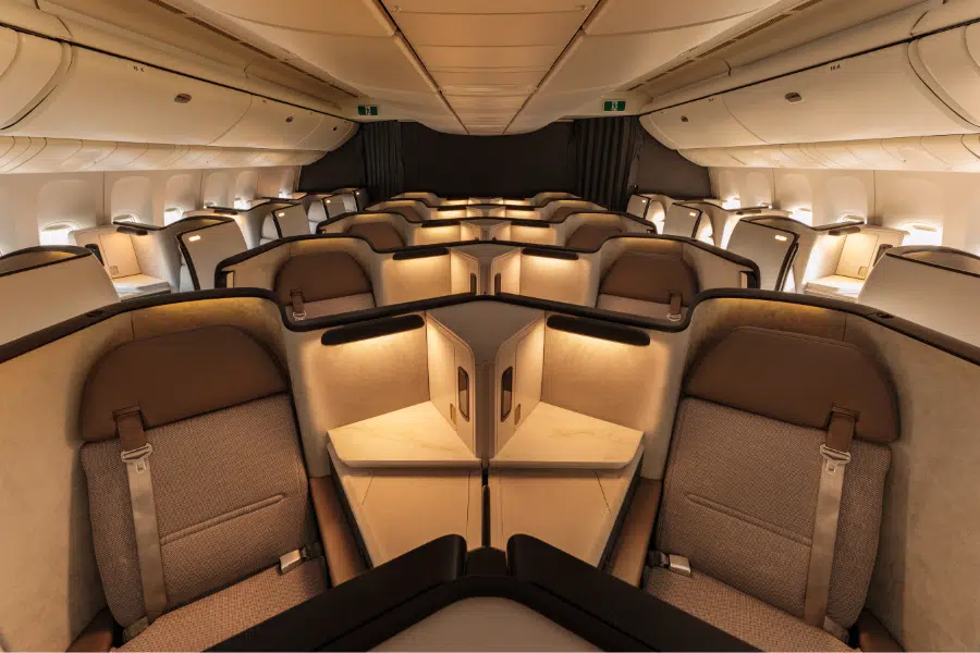 Spacious first-class airplane cabin with wide, beige and brown seats arranged in a 1-2-1 configuration, each with privacy partitions, soft lighting, and large personal space.