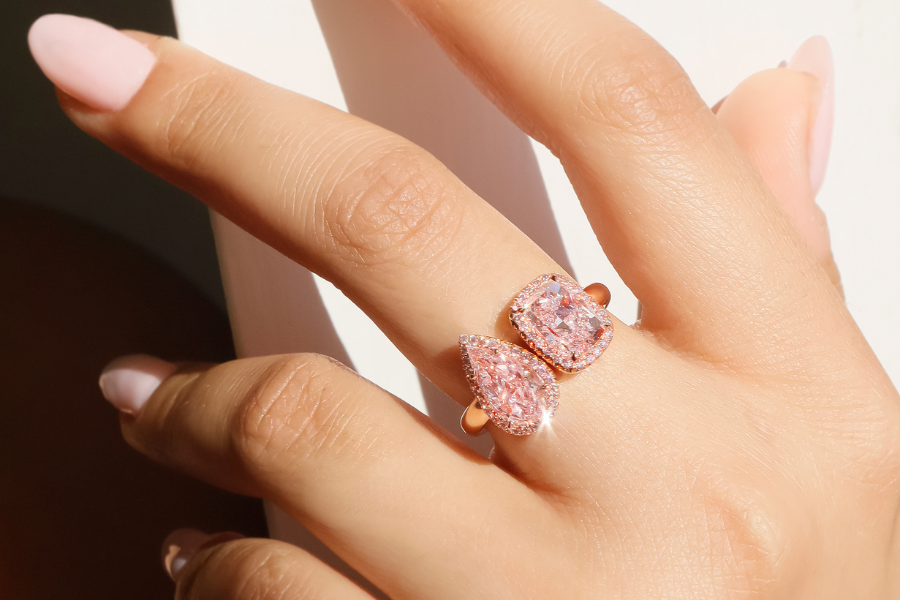 A hand with light pink manicured nails wearing a rose gold ring featuring two large pink gemstones—one pear-shaped and one cushion-cut—sparkling in bright sunlight.