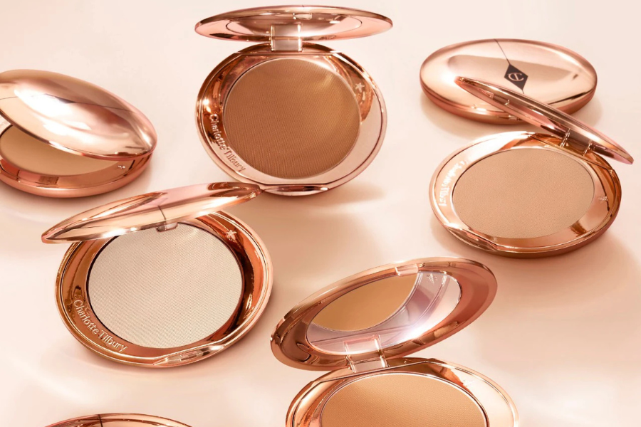 Several open rose gold compact powders with mirrors, displaying different shades of pressed powder makeup, arranged on a light peach surface. The compacts are labeled Charlotte Tilbury.