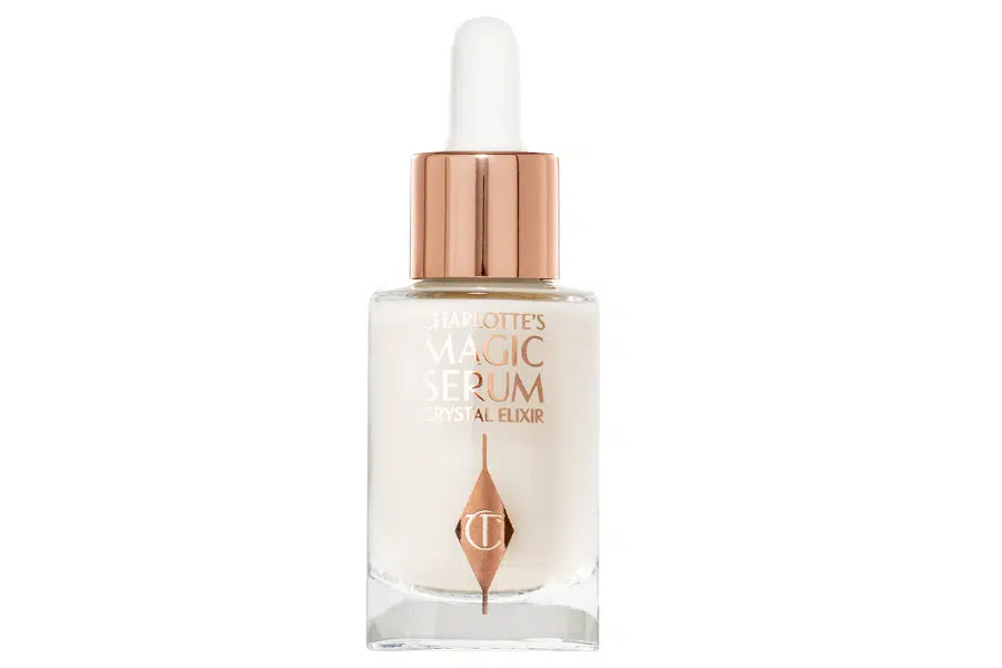 A clear glass dropper bottle with a rose gold cap and white dropper, labeled Charlottes Magic Serum Crystal Elixir with a logo on the front. The liquid inside appears milky white.