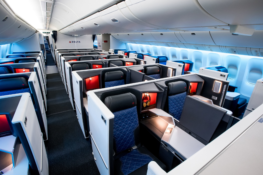 The image shows the interior of a Delta Air Lines aircraft, featuring a modern business class cabin with spacious, private seats arranged in a staggered layout, each with individual screens and sleek blue and white accents.