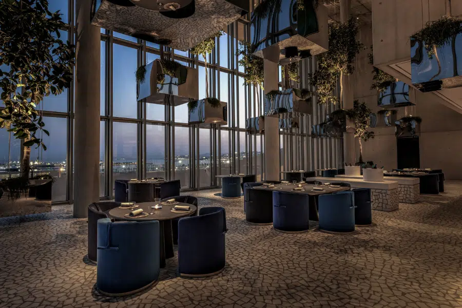 A modern, elegant restaurant with floor-to-ceiling windows, blue and black chairs, round tables set for dining, stone-tiled flooring, and lush greenery hanging from the ceiling. City lights are visible outside at dusk.
