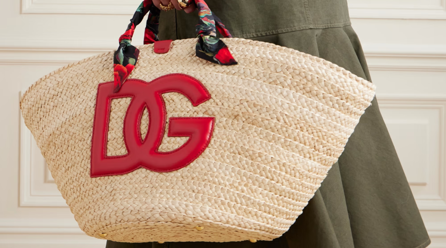 A person holds a woven straw handbag with large red “DG” letters and a colorful scarf wrapped around the handle, wearing an olive green outfit.