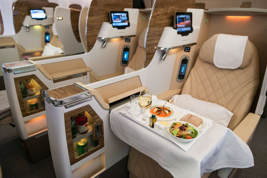A luxury airline business class seat with a beige leather recliner, TV screen, drinks shelf, and a tray table set with a gourmet meal, bread, salad, wine, and water. The area looks modern and spacious.