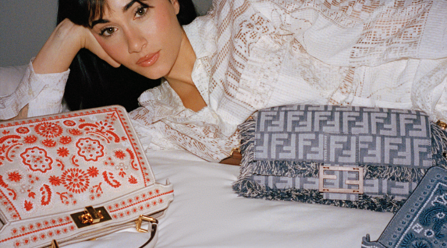 A woman in a white lace blouse lies on a bed beside two designer handbags: one with red embroidery and the other with a blue and gray Fendi logo pattern.