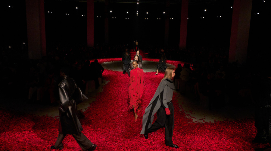 Models walk down a runway covered in red petals, wearing dramatic red and dark outfits, with dim lighting and an audience seated on both sides in a spacious indoor venue.